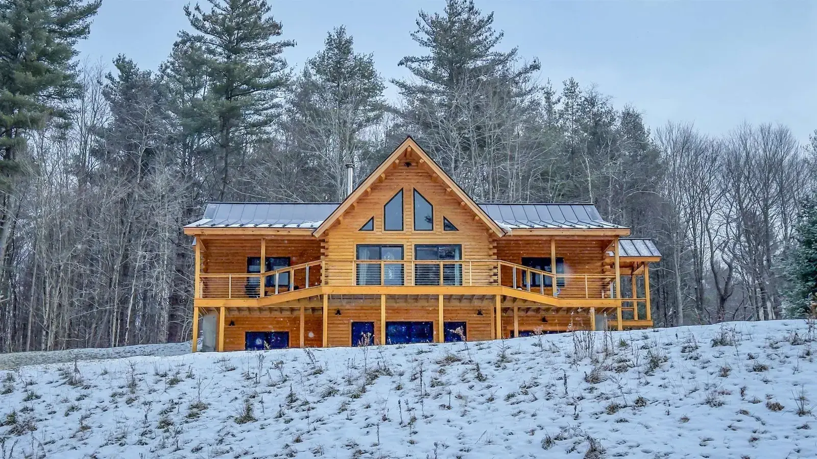 Cedar log home with a covered deck and large gable windows on a wooded winter hillside