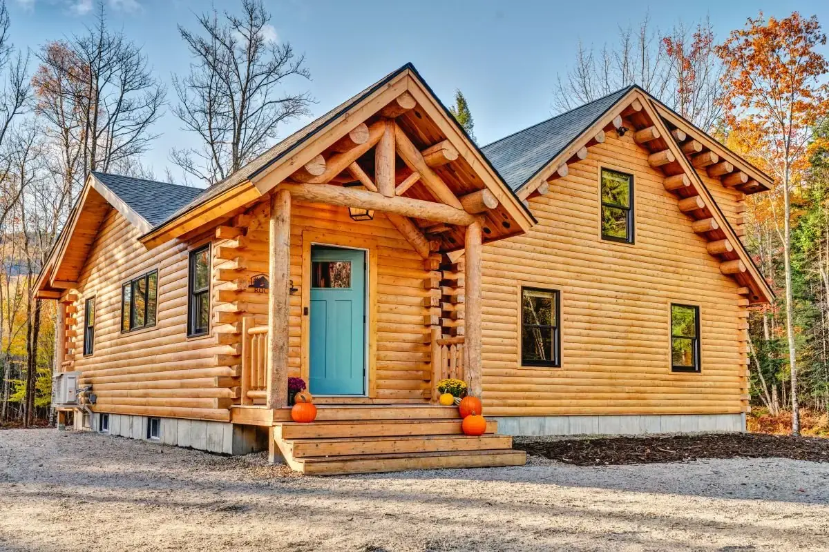 Cedar log home with a timber king-post porch, teal front door, round log corners, and black-trimmed windows among autumn trees