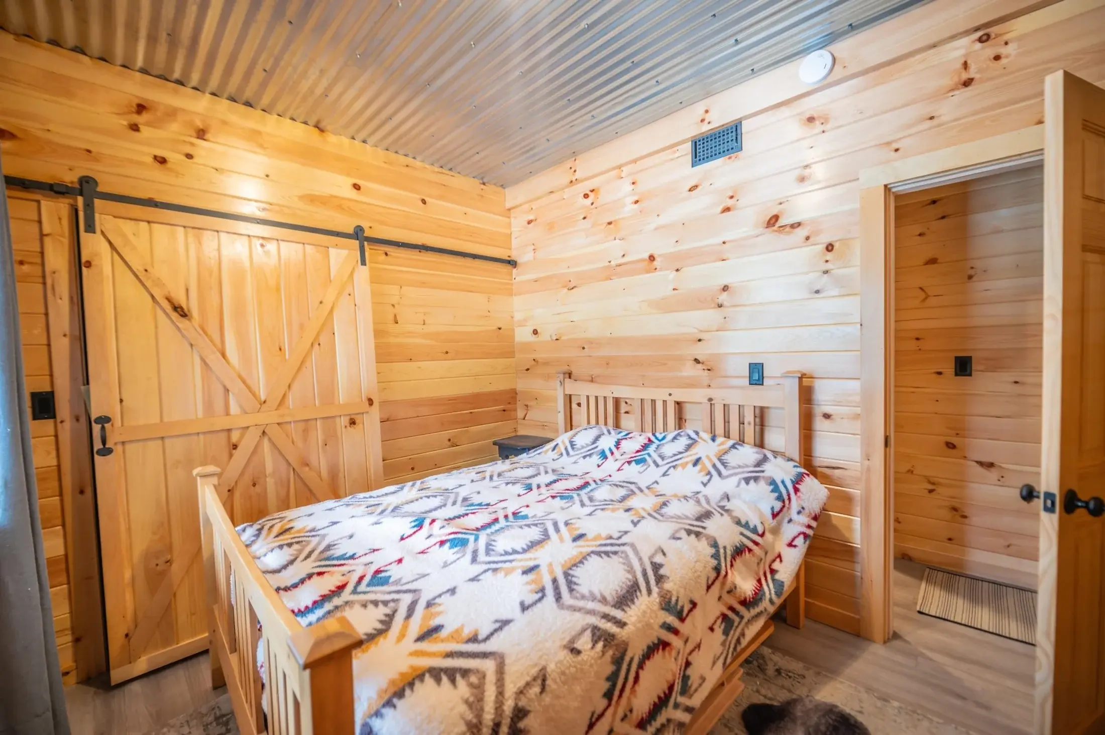 This bedroom continues the rustic industrial style with a sliding barn door on sturdy black hardware. A colorful quilt adds a layer of soft texture, creating a harmonious and restful space. 