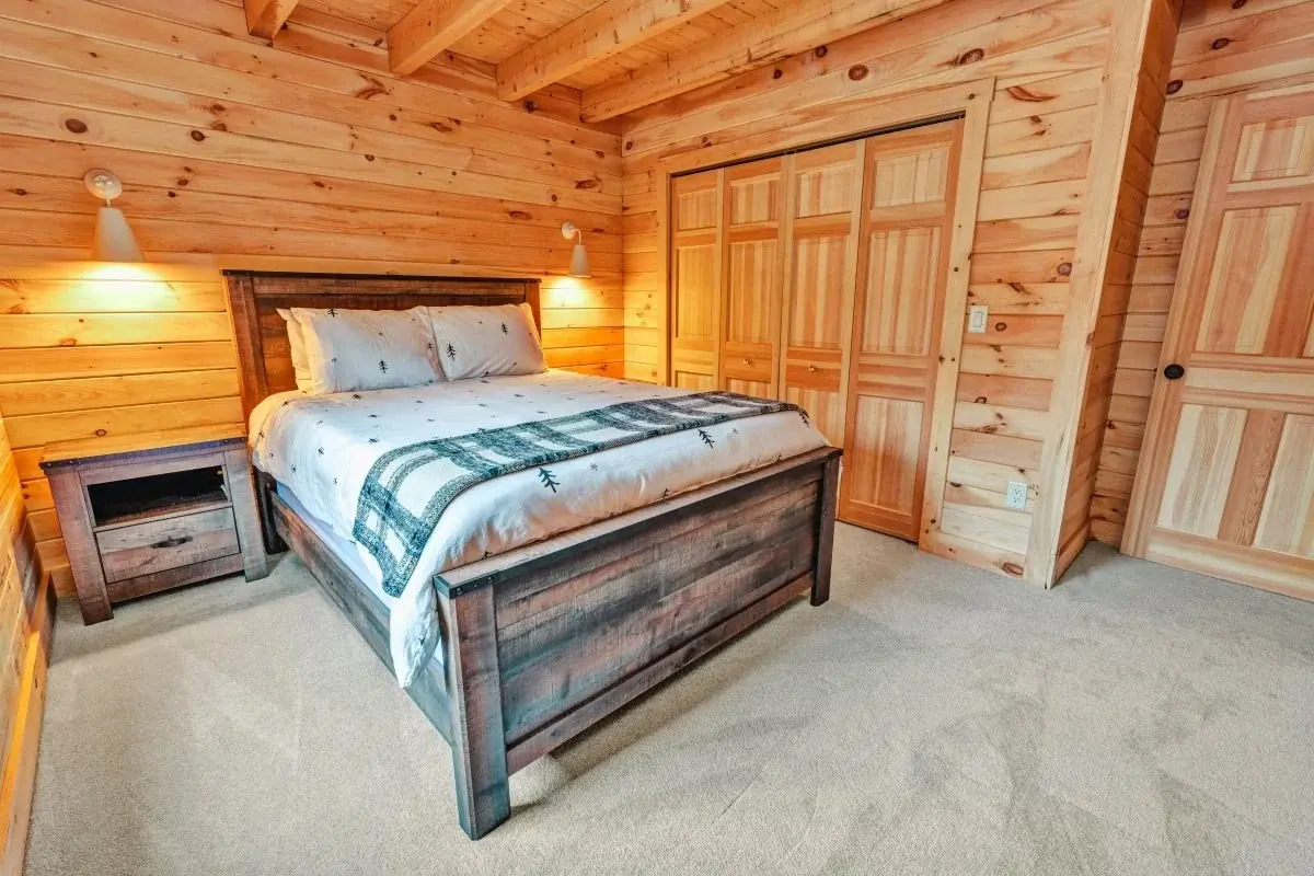 A cozy, rustic bedroom offers a serene retreat. The plush light carpet and soft bedding are a soft contrast to the solid wood of the walls, ceiling, and furniture. Simple matte white wall sconces add a touch of modernity while still coordinating with