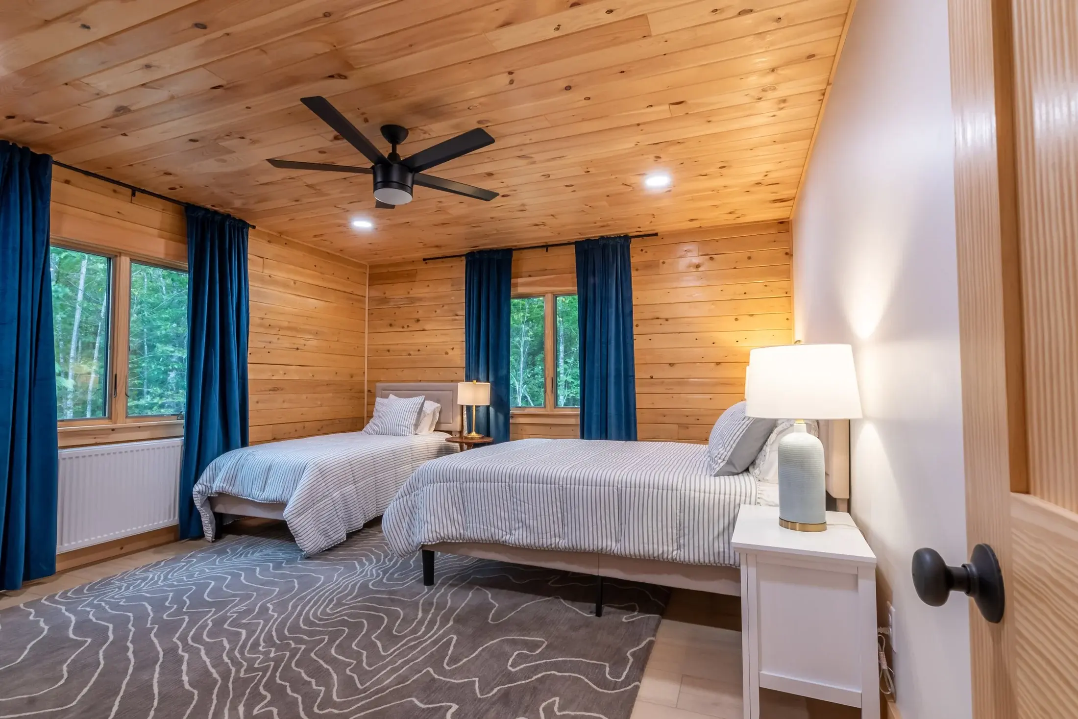 A fresh white accent wall and simple furnishings keep this tidy bedroom classic and comfortable. The rustic wooden walls and light wood floor are balanced by a modern area rug, crisp striped bedding, and sleek black hardware. 