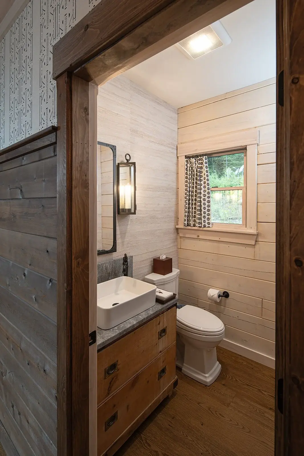 In the first floor powder room, a vintage-inspired vanity cabinet is topped with a modern rectangular vessel sink for a balanced style and rugged practicality. The black and white curtains reflect the geometric patterned wallpaper in the hallway, ano