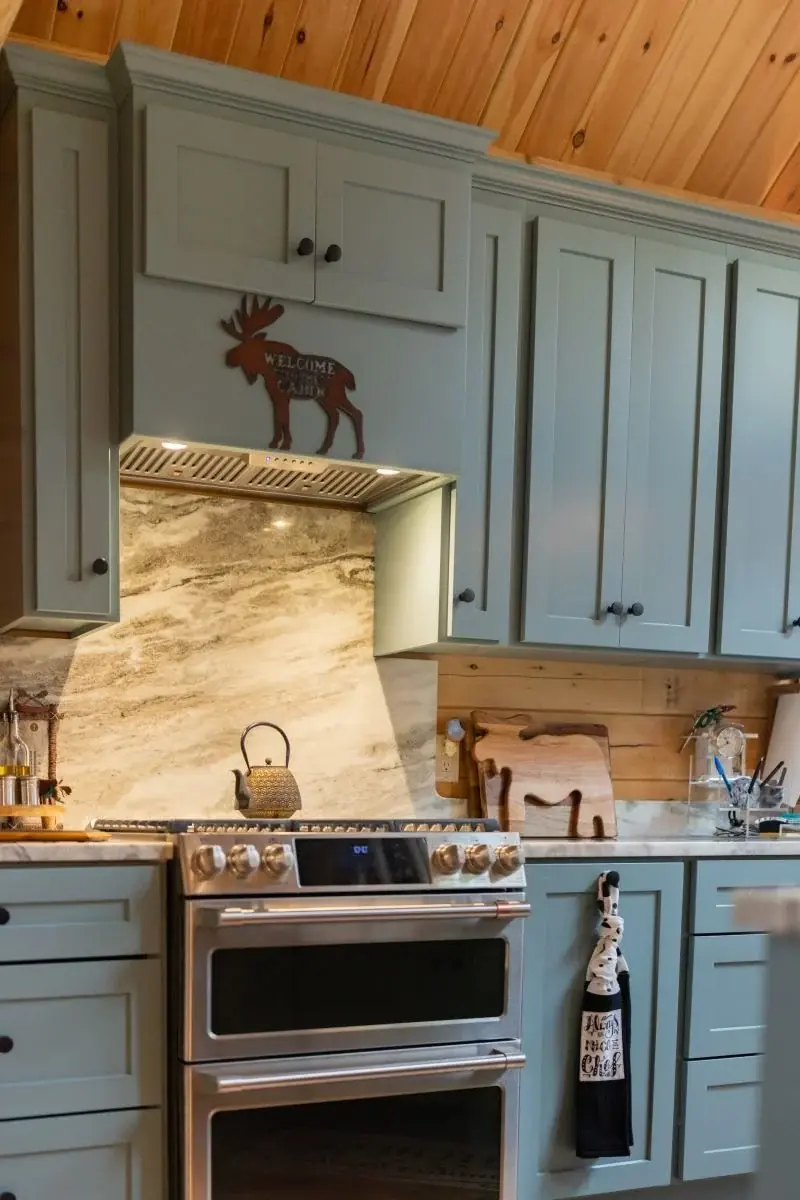 Gray and white quartzite countertops transition seamlessly into a high backsplash above the stove. Elegantly paired with muted teal cabinetry, it’s the perfect complement to the golden wood tones. 