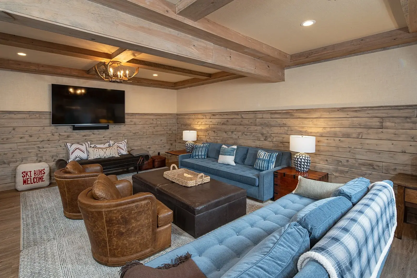 In the family room, warm browns and casual blues combine with rustic wood paneling and exposed beams to create a relaxed and comfortable space. Layers of light like an antler ceiling pendant, recessed puck lights, and stylish table lamps add to the a