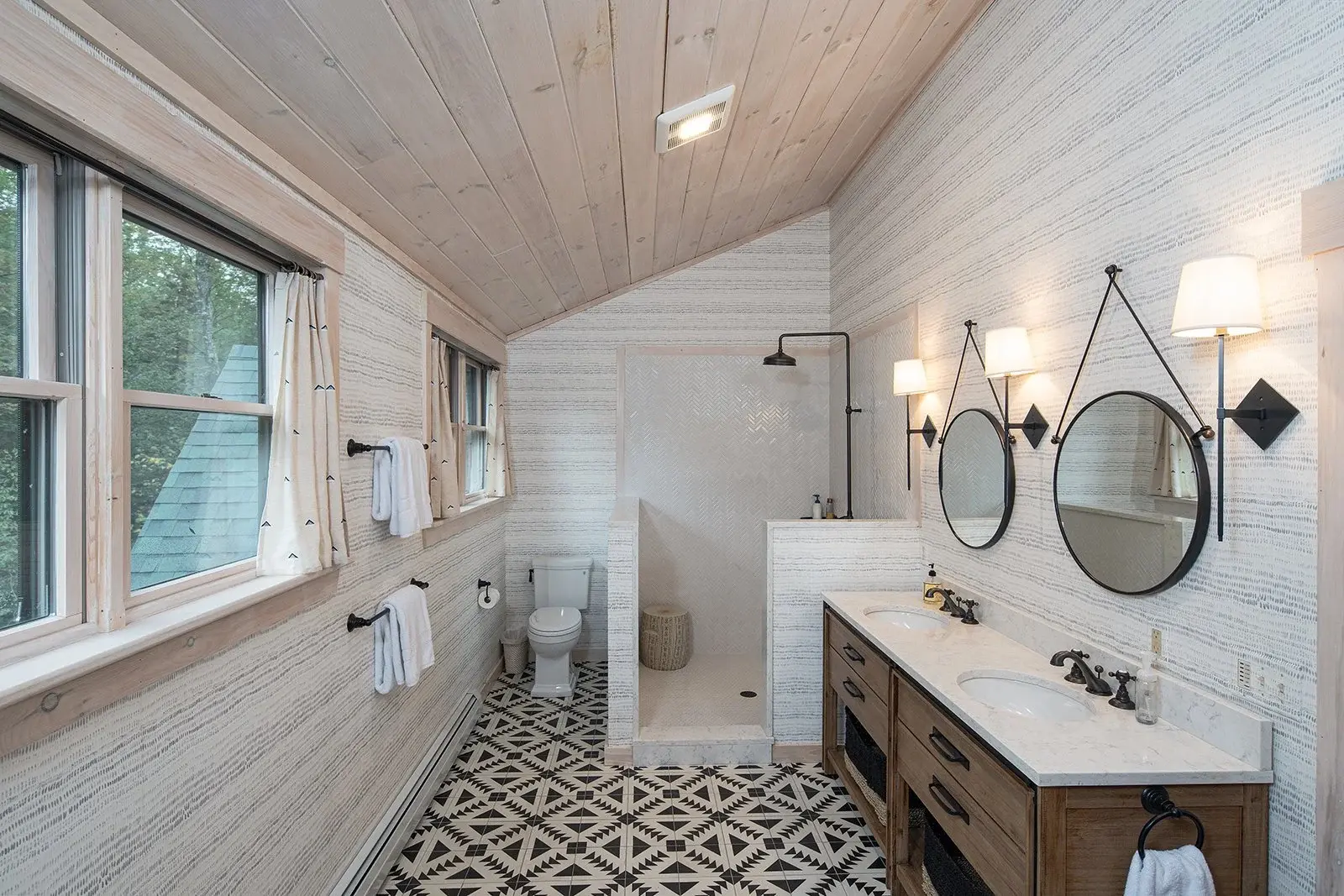 Pickled white ceiling boards transition effortlessly to a simple patterned wallpaper, letting the black and white geometric floor panels stand as the focal point. A natural wood vanity and simple black fixtures are consistent with the sturdy, underst